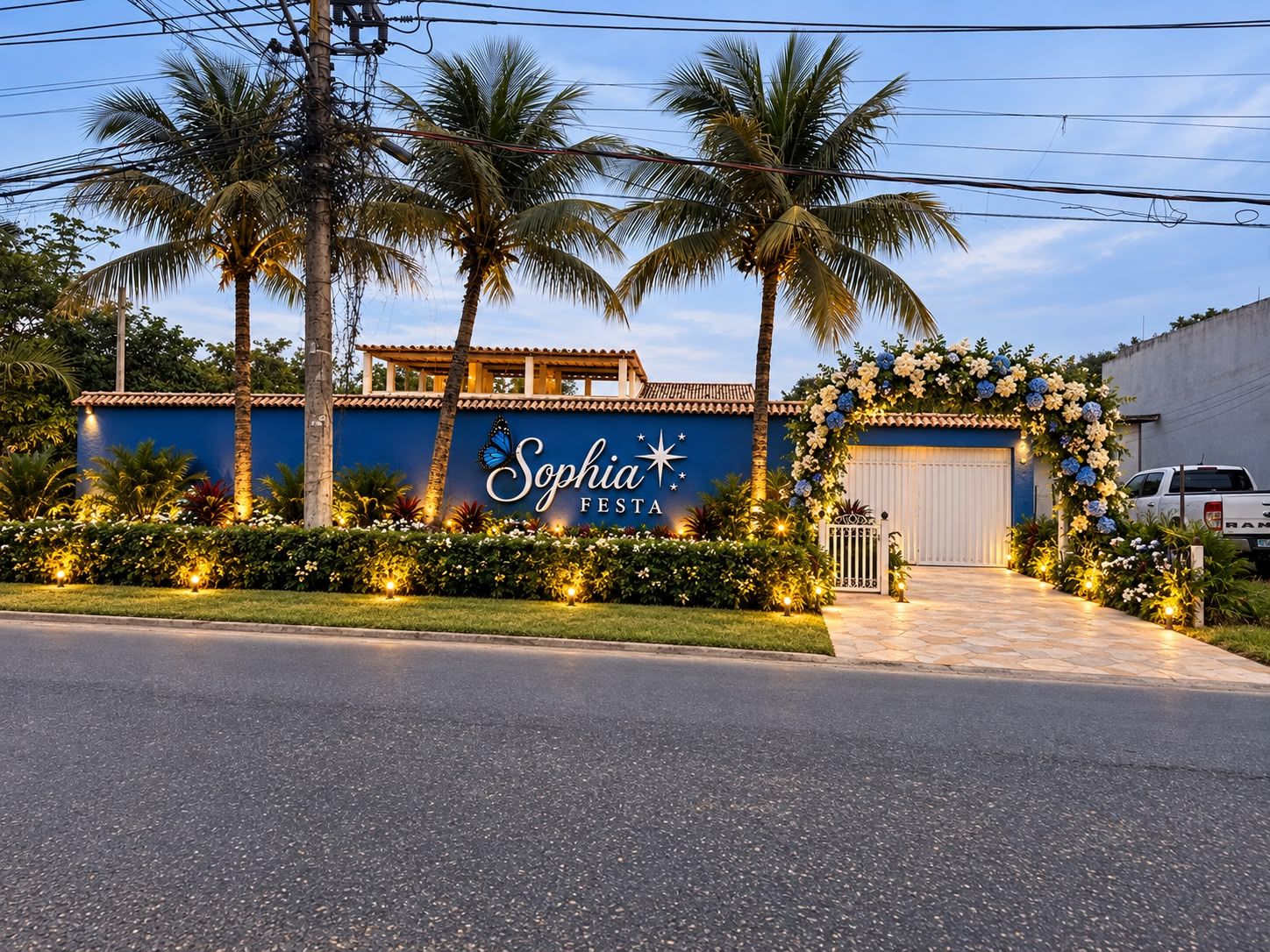 Fachada ao entardecer, com muro azul e letreiro, palmeiras e arco de flores no portão